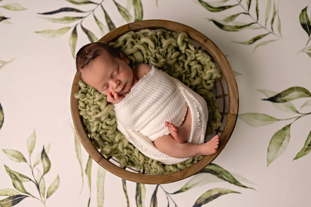 Newborn sleeping peacefully in wooden bowl with green fabric and botanical leaf backdrop.