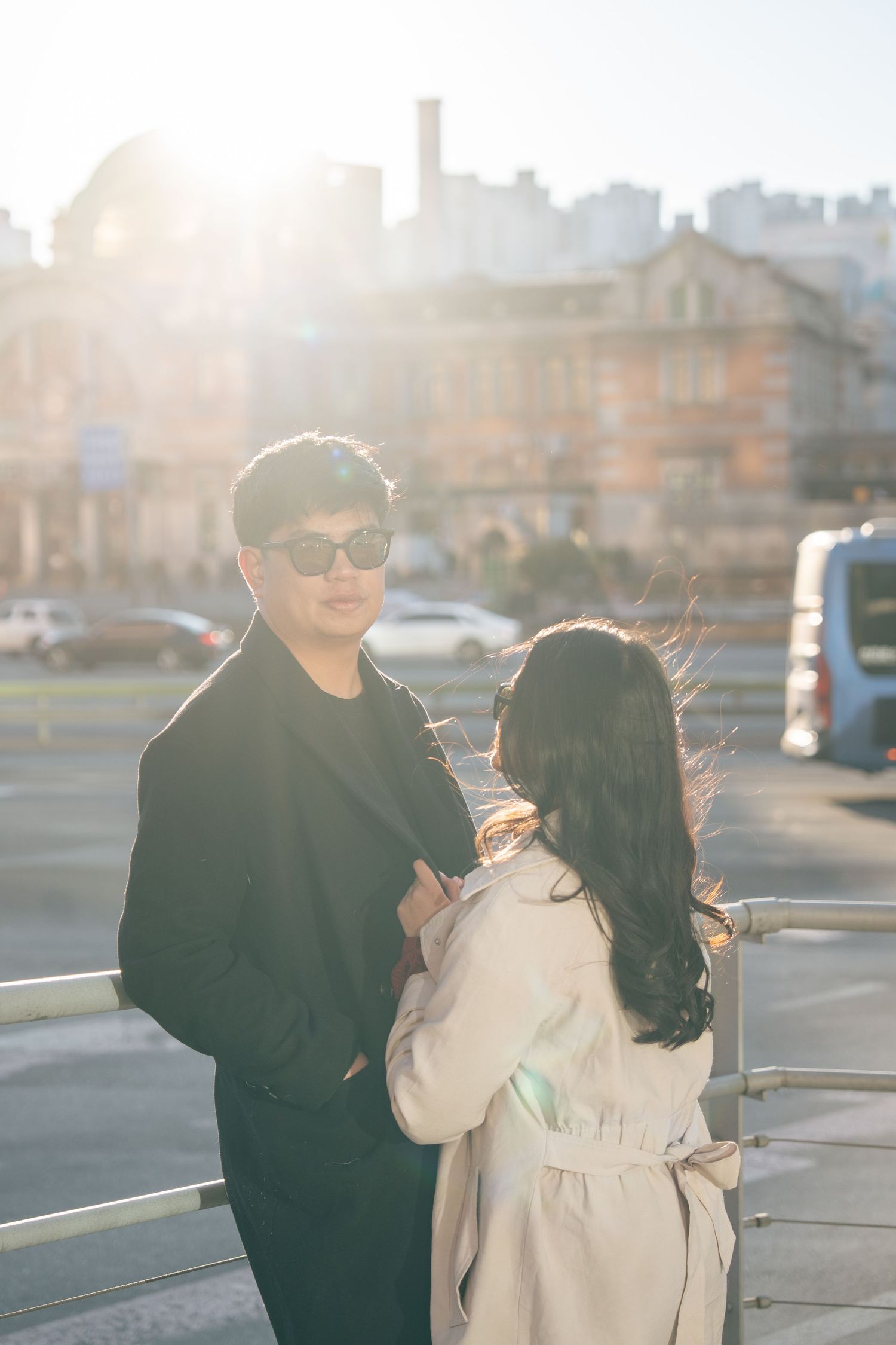 A couple in stylish coats stands together on a rooftop during golden hour with a cityscape in the background.