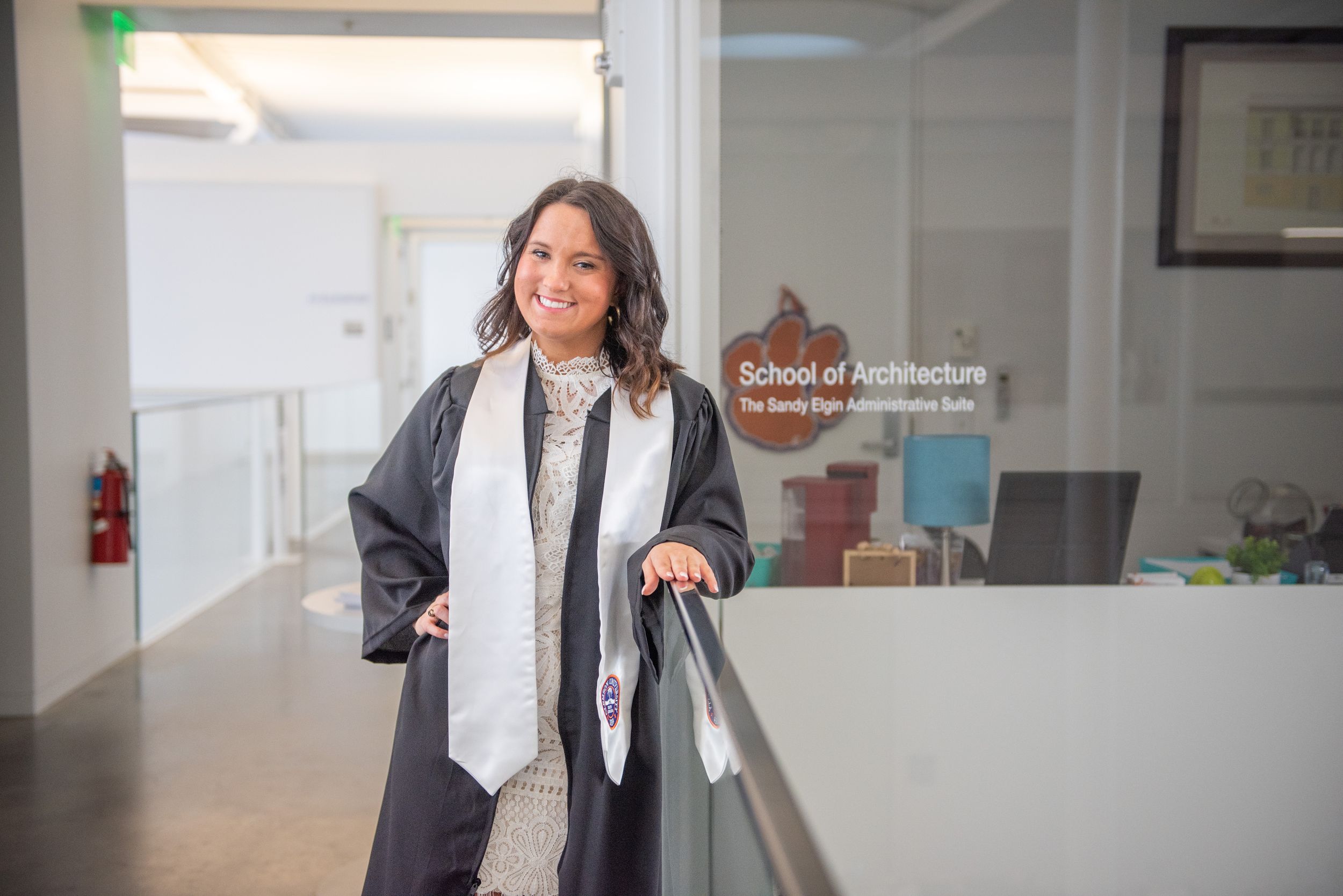 Professional in a graduation gown and white stole stands smiling in a bright hallway with glass walls.