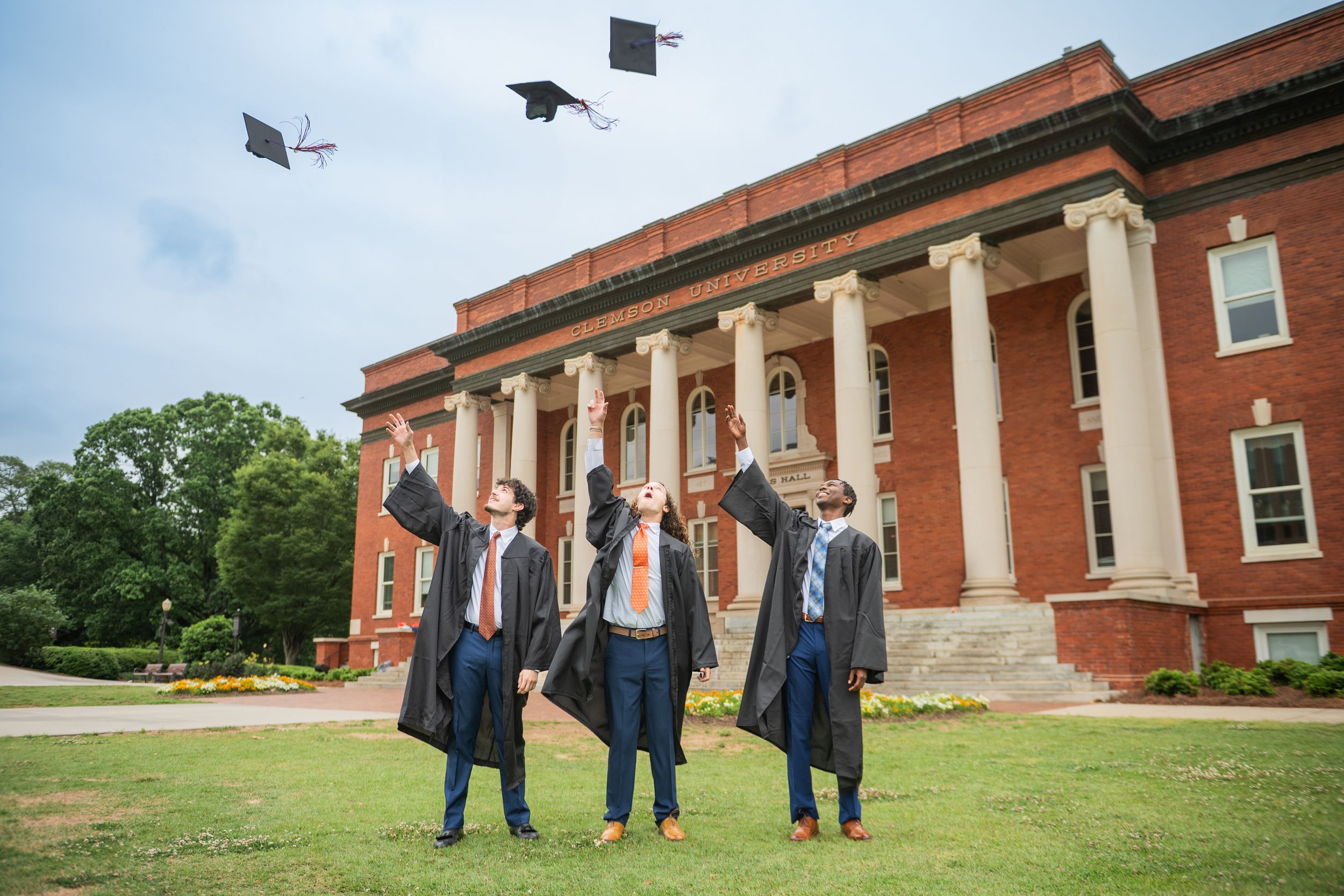 Graduates in blue caps and gowns walk together in front of a red brick building on a sunny day.