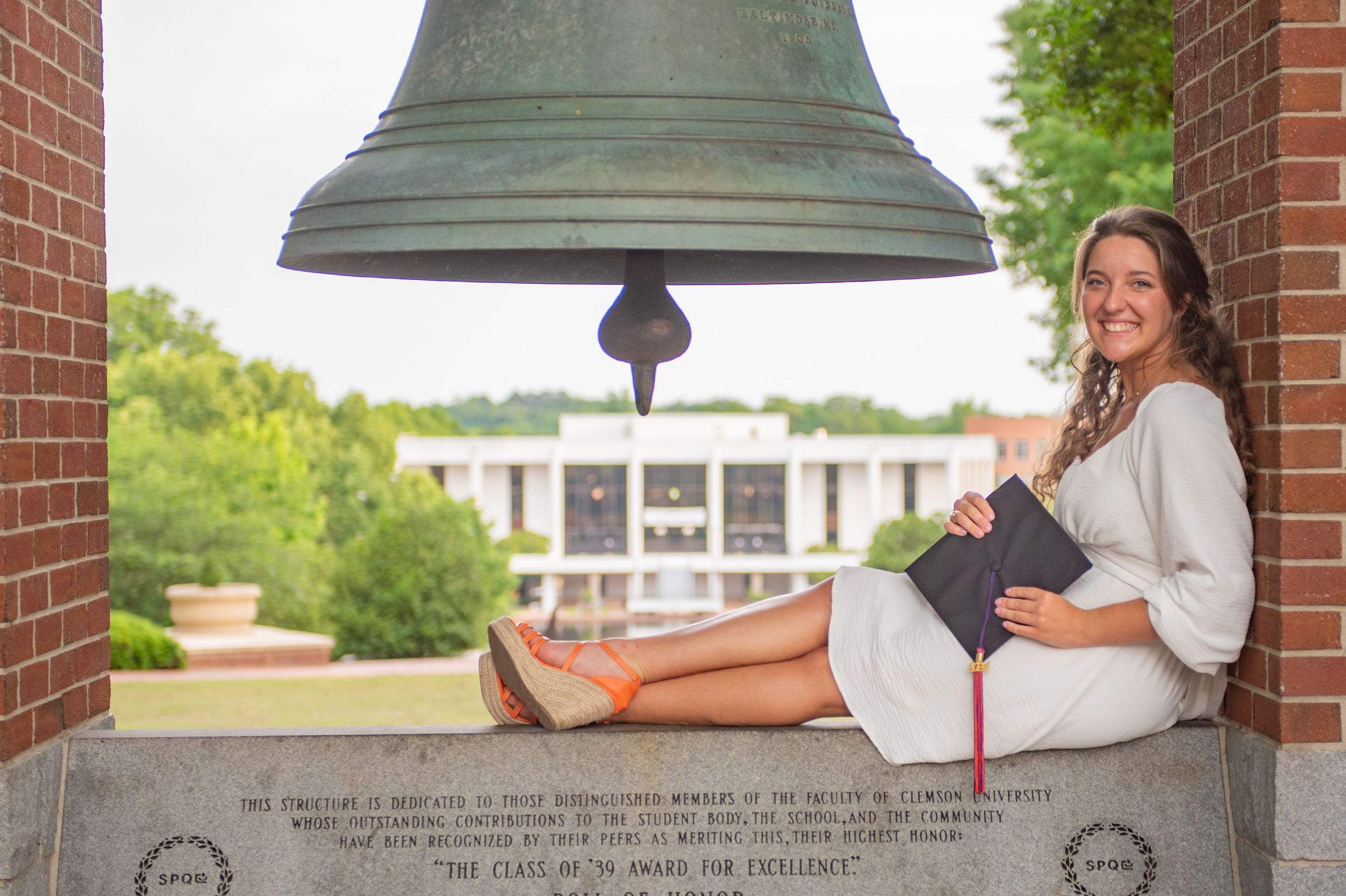 A couple sits together on a bench under a large bell at a college campus entrance.