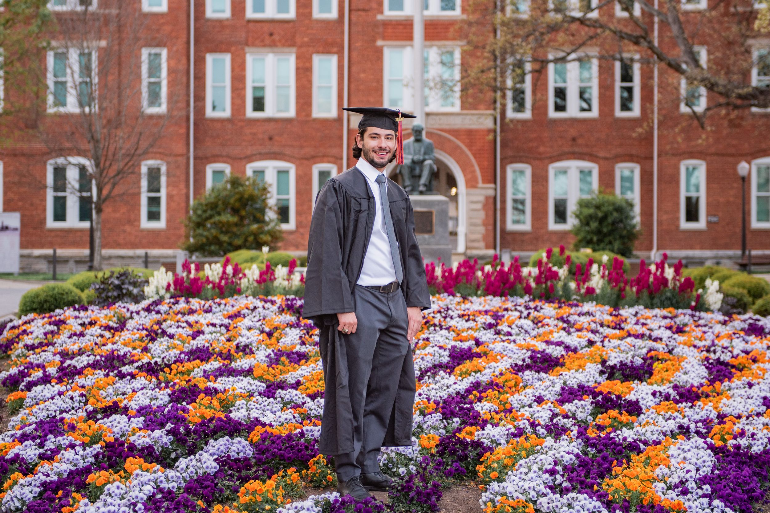 Graduate stands in a suit and cap among vibrant purple and orange flowers blooming in front of a brick university building.