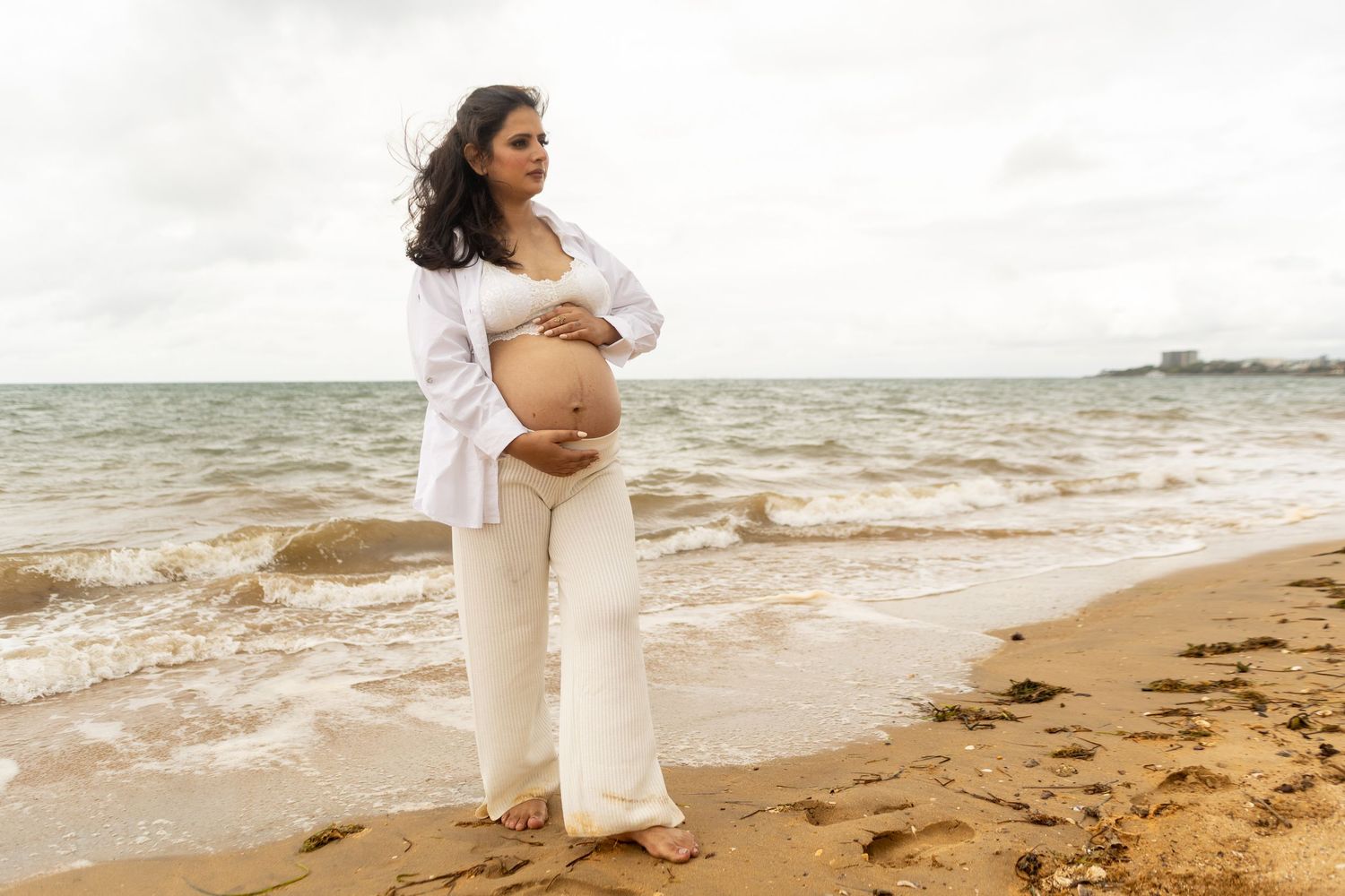 A person in a white shirt and pants walks along a sandy Brisbane beach at sunset while holding their pregnancy bump.