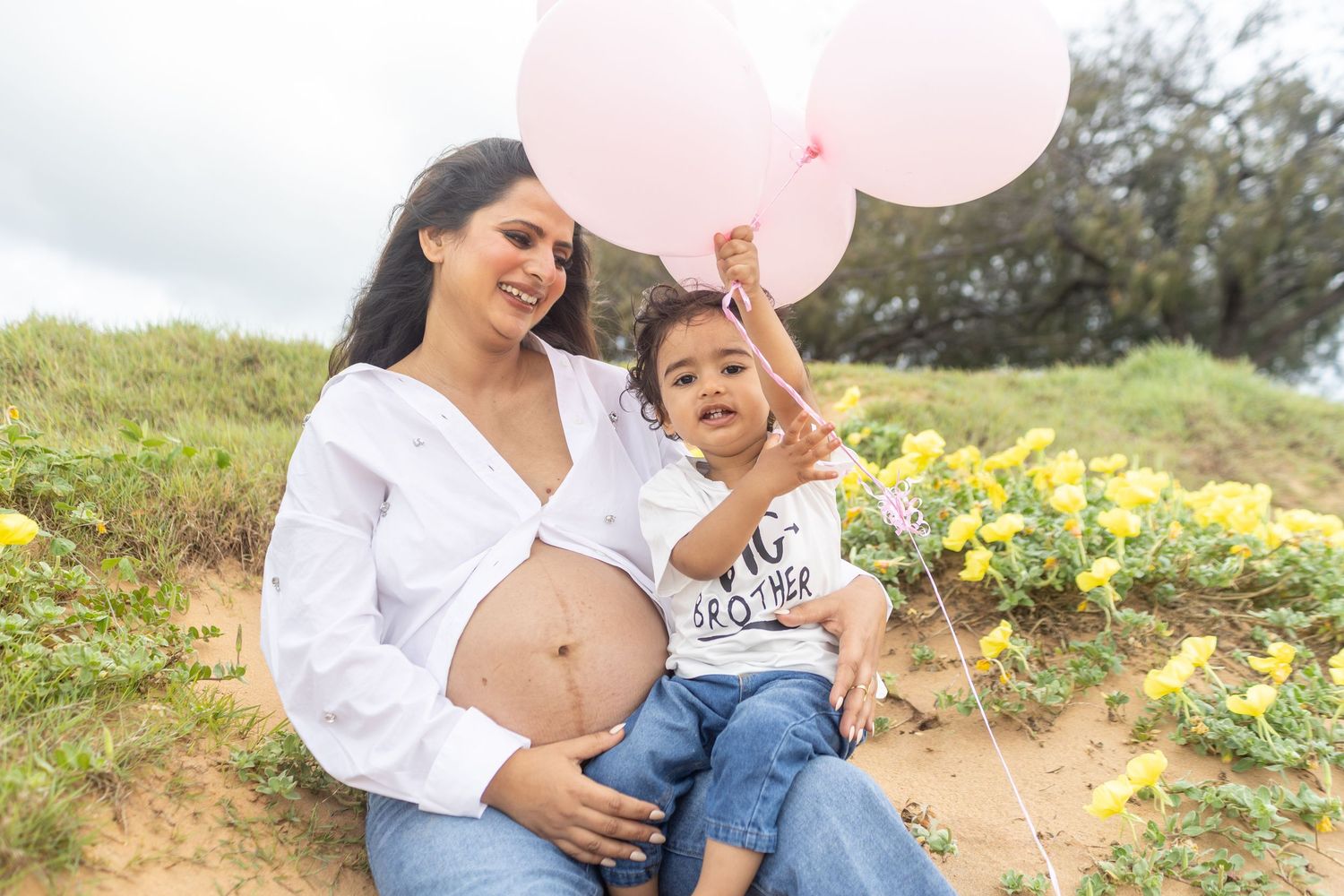 A family shares joyful moments on a Brisbane beach with yellow wildflowers while holding pink balloons on a sunny day.