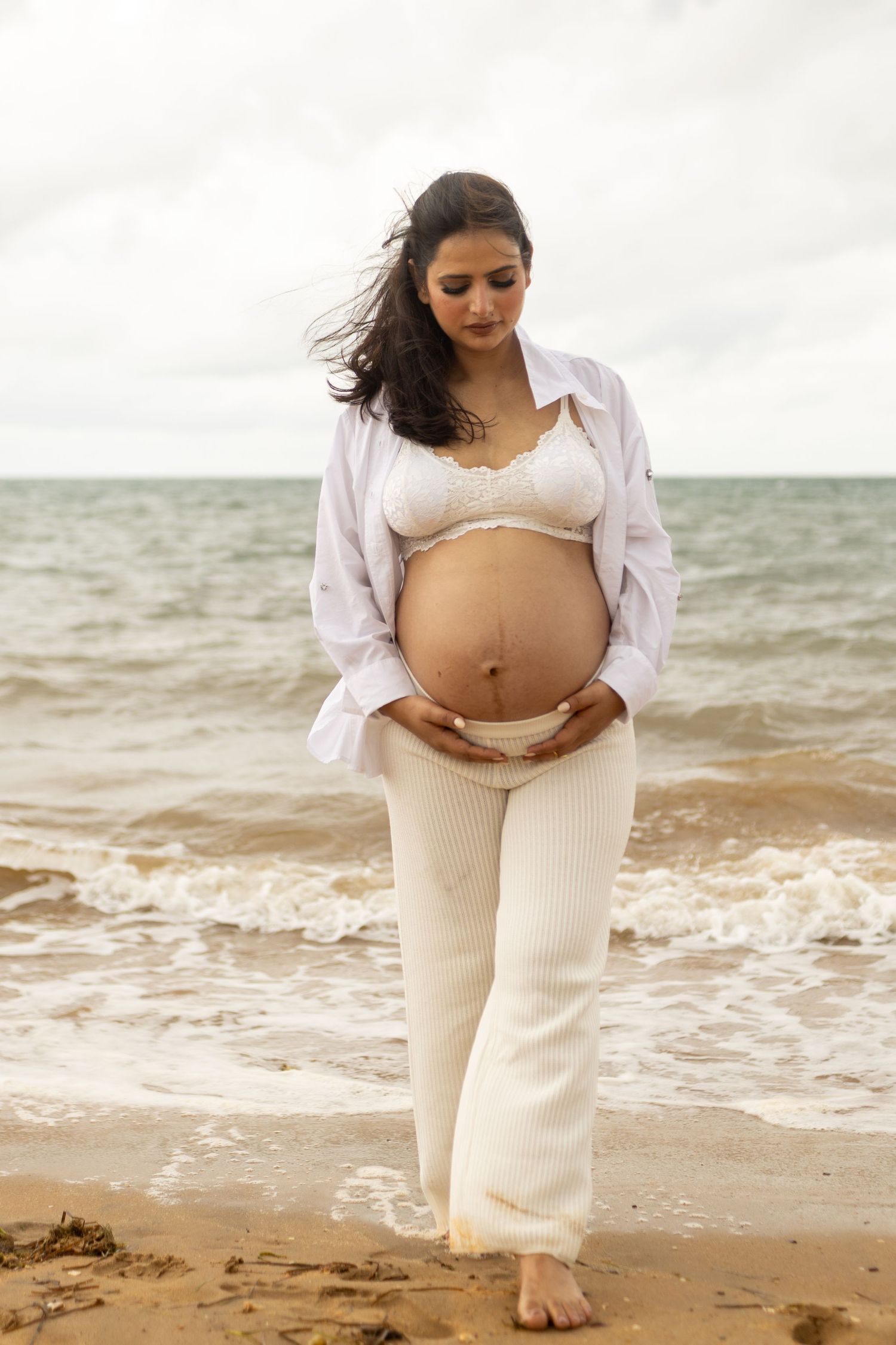 A maternity photo session on a Brisbane beach shows a pregnant woman in white clothing gazing at her belly by the ocean waves.
