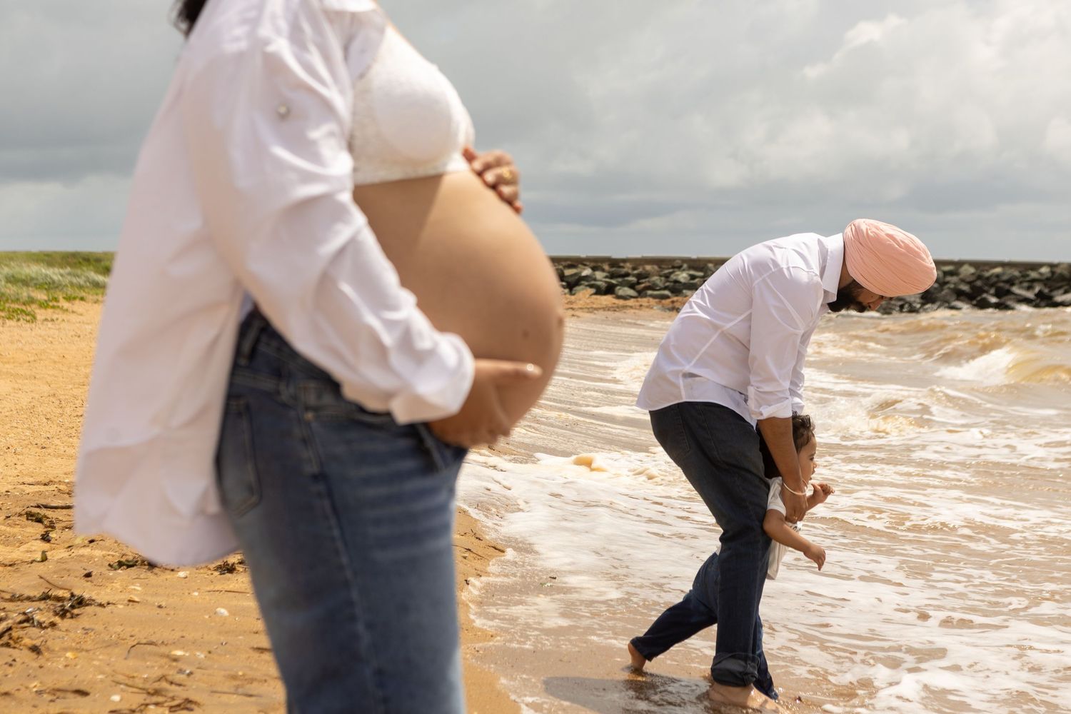 A pregnant woman in a white shirt and jeans walks along a sandy Brisbane beach with waves crashing on the shore.