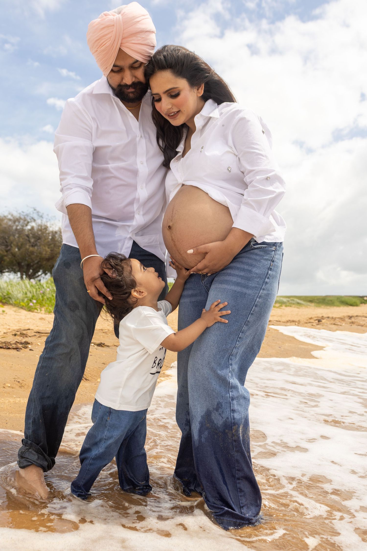 Couple in white shirts and jeans share intimate moments on a Brisbane beach while expecting a baby.