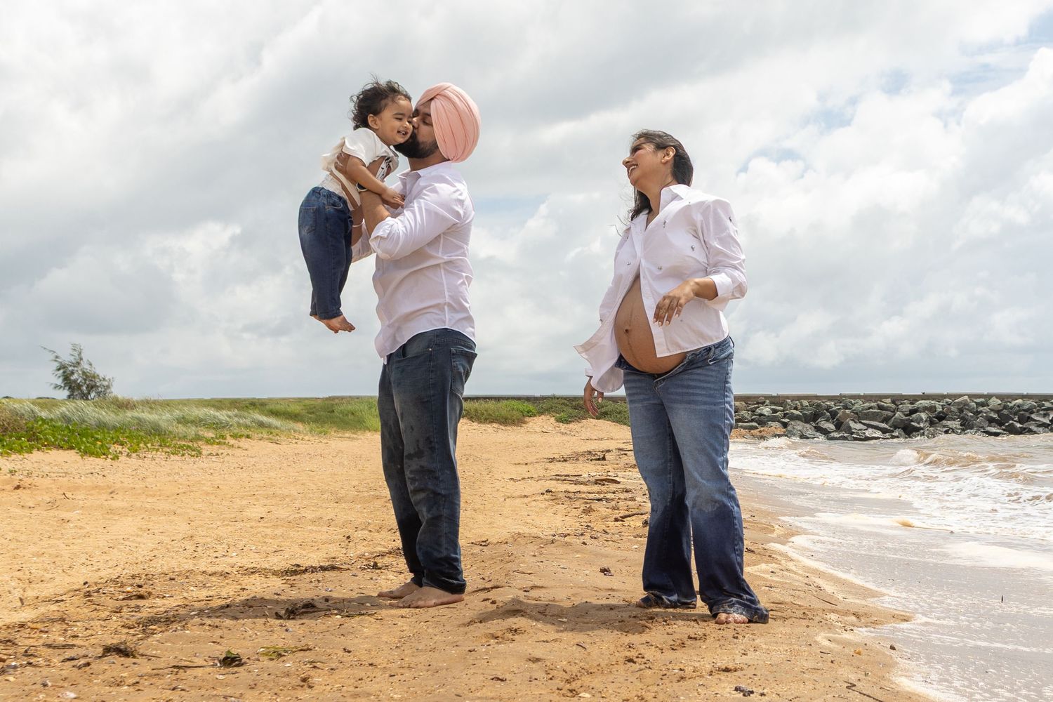 A joyful family moment captured on a sandy Brisbane beach with cloudy skies and ocean waves in the background.