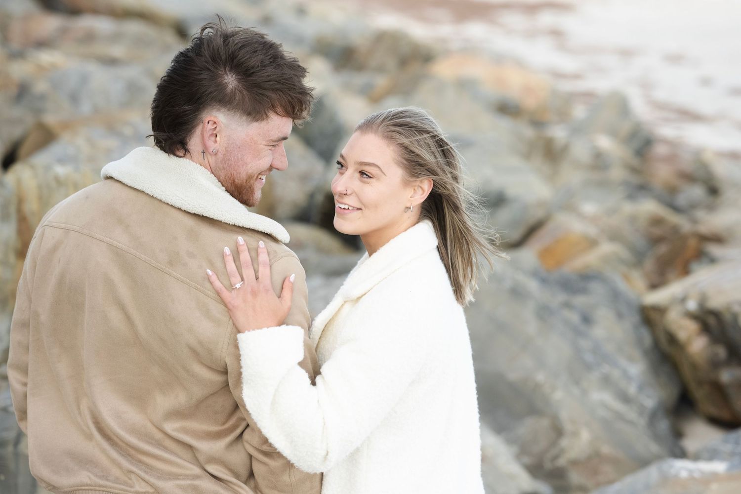Couple embracing and smiling near rocky beach, woman in white coat with engagement ring visible.