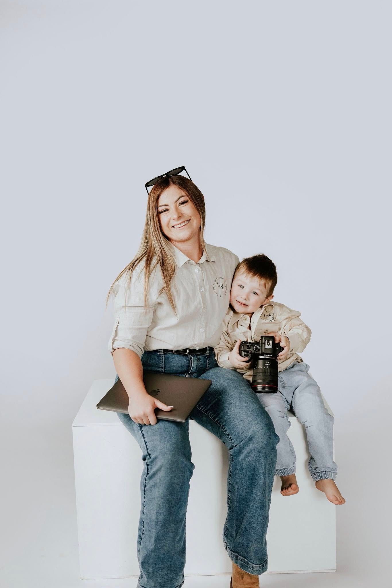 Smiling female photographer sitting with young son holding camera on white studio cube.