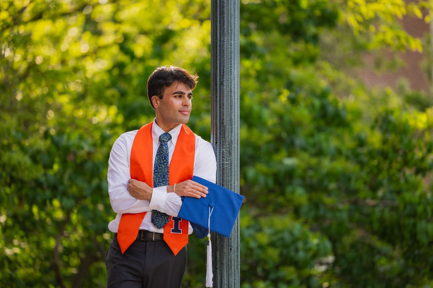 Graduating student at UIUC leaning against a pole and posing with a graduation cap at Foellinger