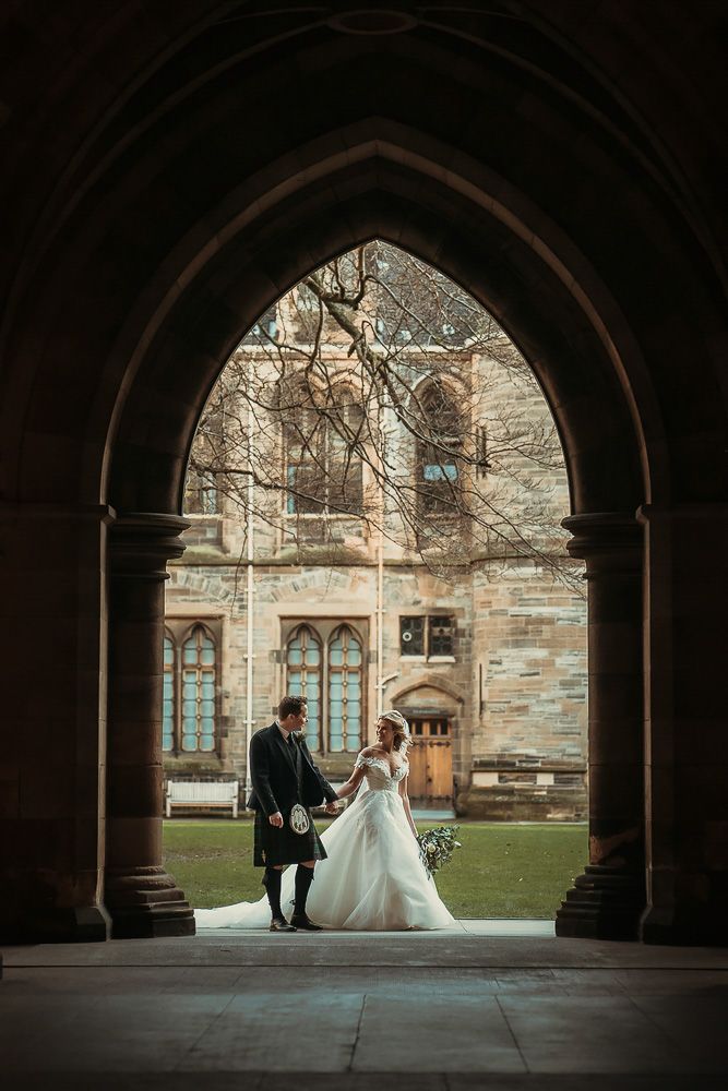 Wedding couple posed under gothic stone archway at historic building.
