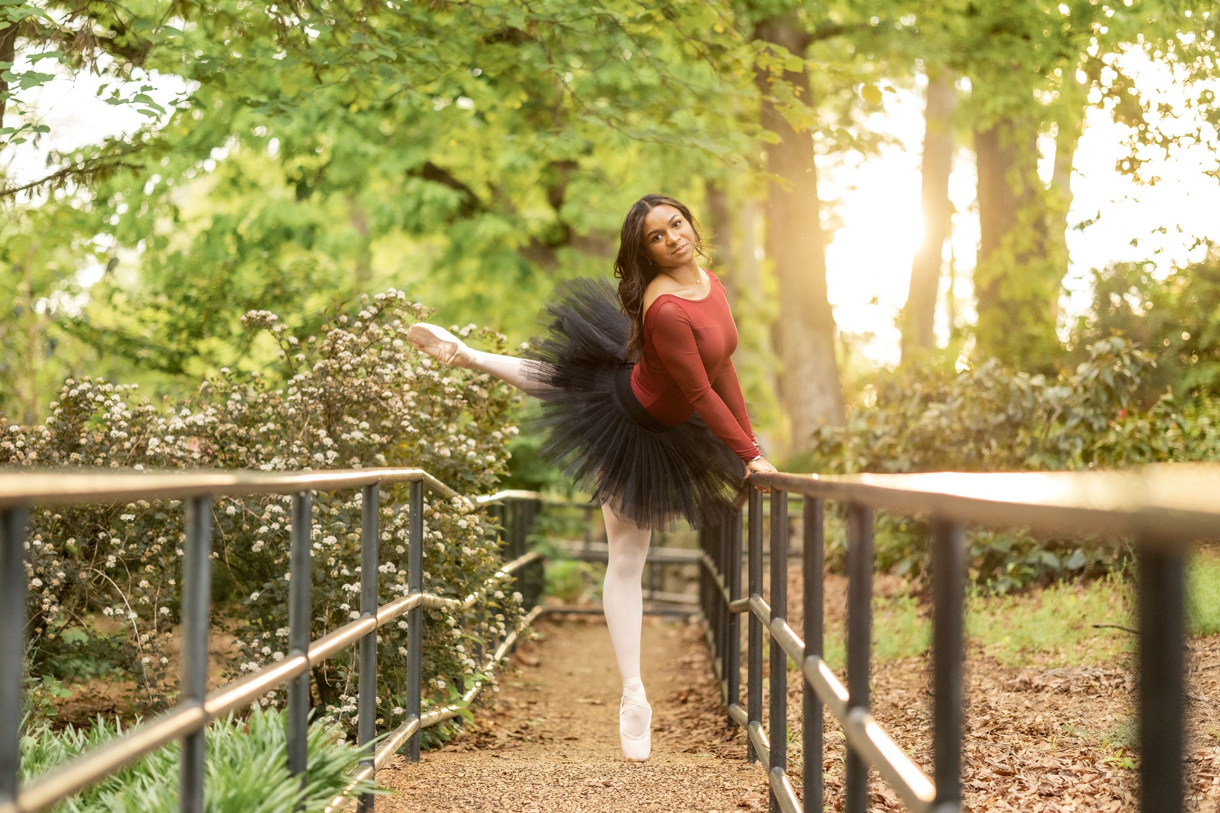 Dancer in a black tutu performing graceful movements on a wooden bridge surrounded by lush green trees.
