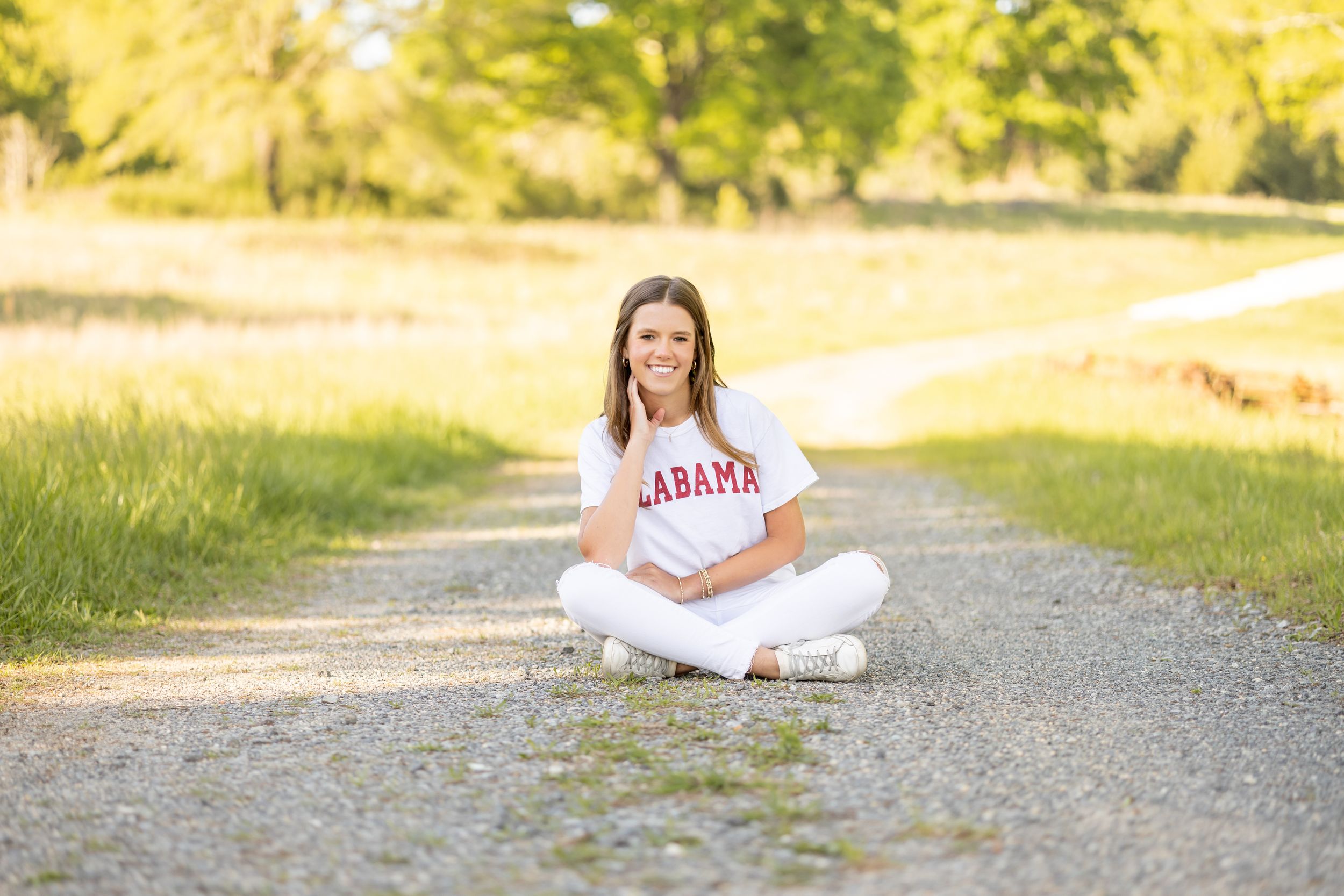 A person in white clothing sits on a gravel path surrounded by lush greenery and warm sunlight.