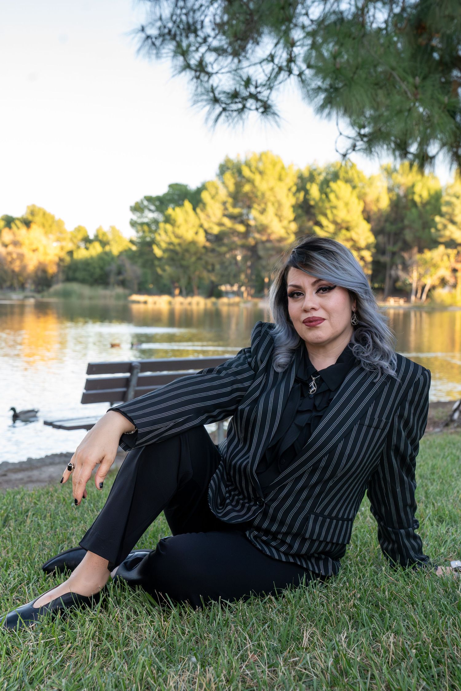 Professional outdoor photo shoot by a lake featuring someone in a black and white striped blazer reclining on grass.