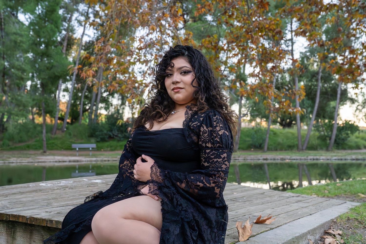A person in a black lace dress poses on wooden steps surrounded by autumn trees in a park setting.