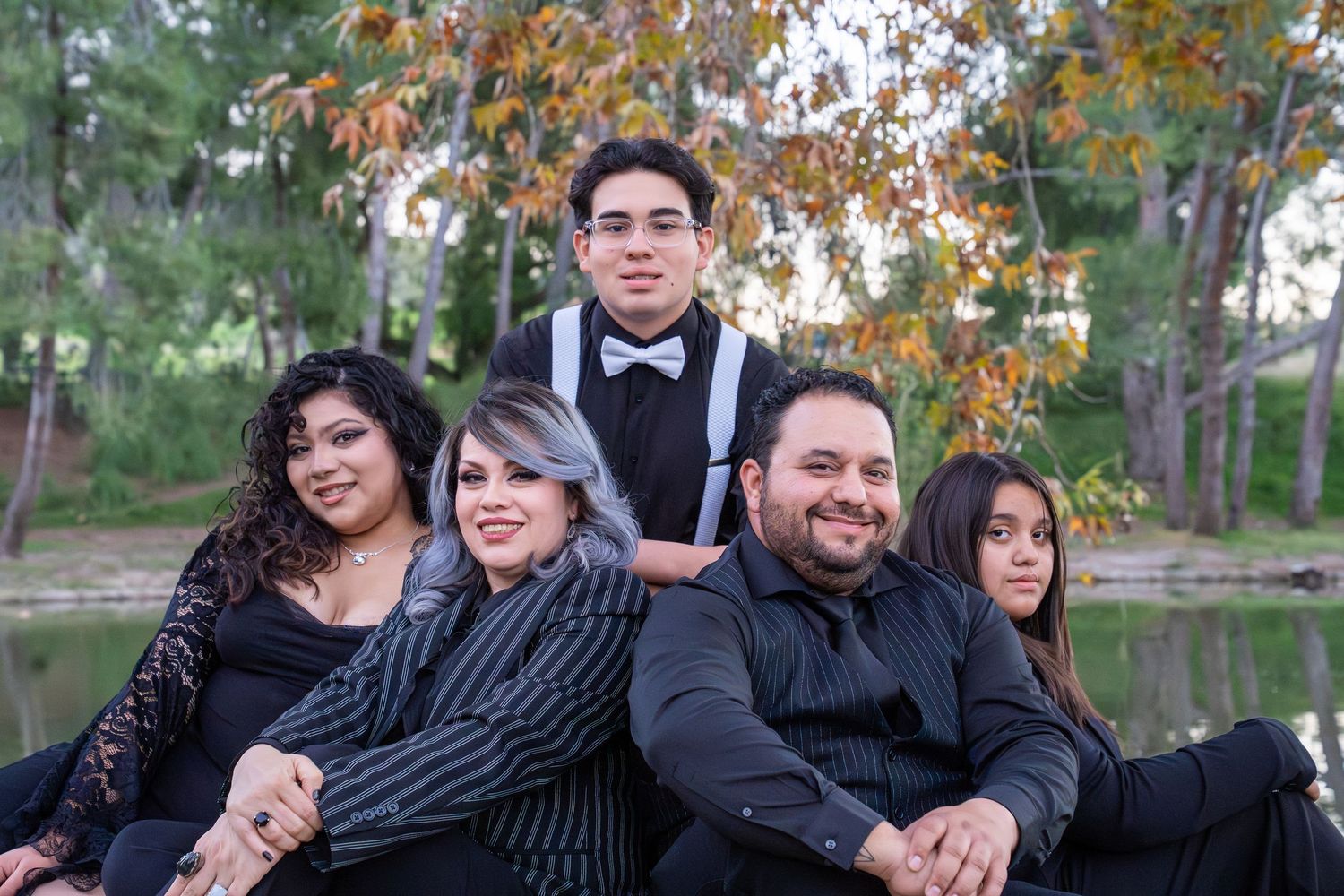A family in formal attire poses together outdoors during autumn with fall foliage and a lake in the background.
