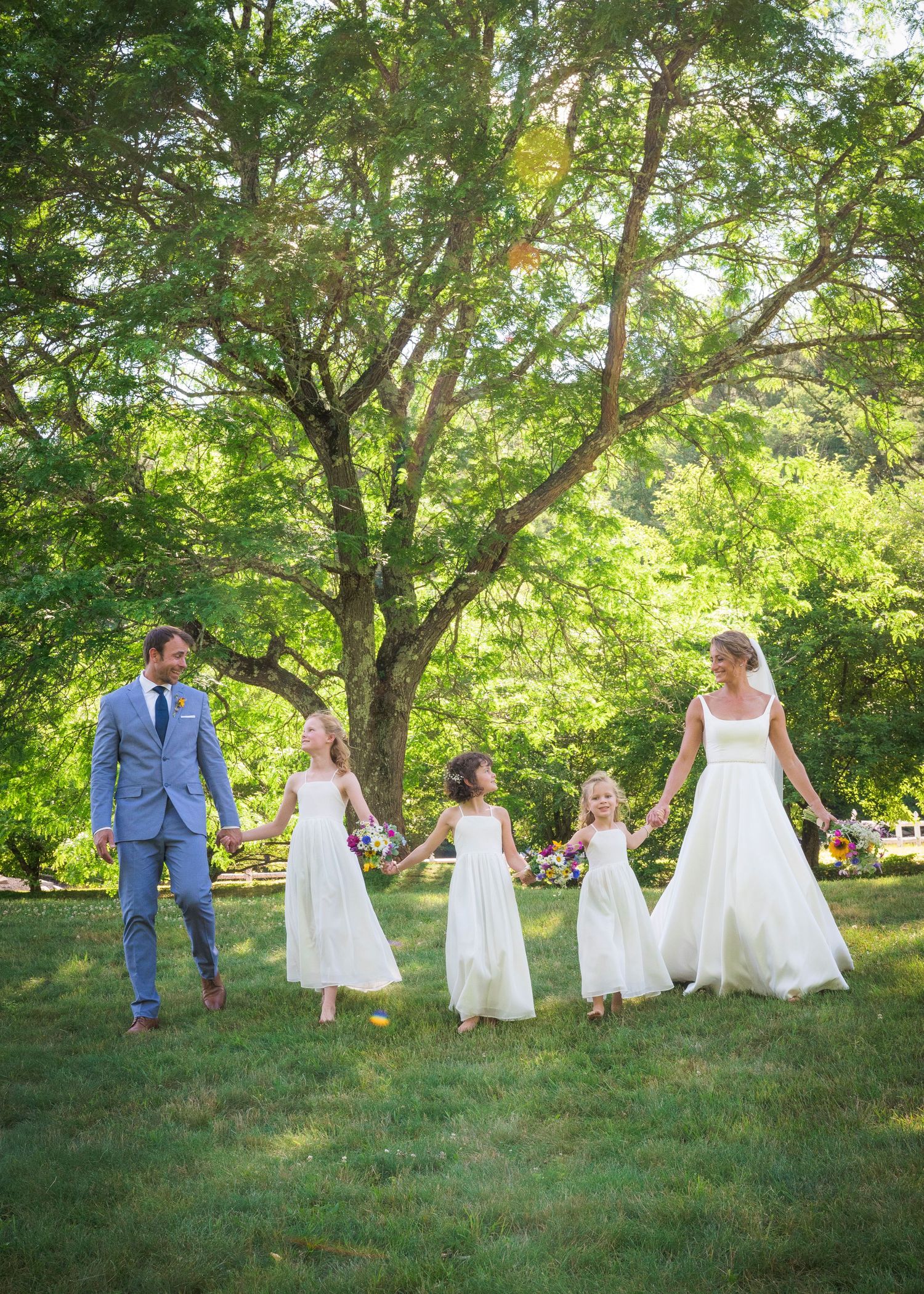 A wedding party stands together under a large tree on a sunny summer day in a garden setting.