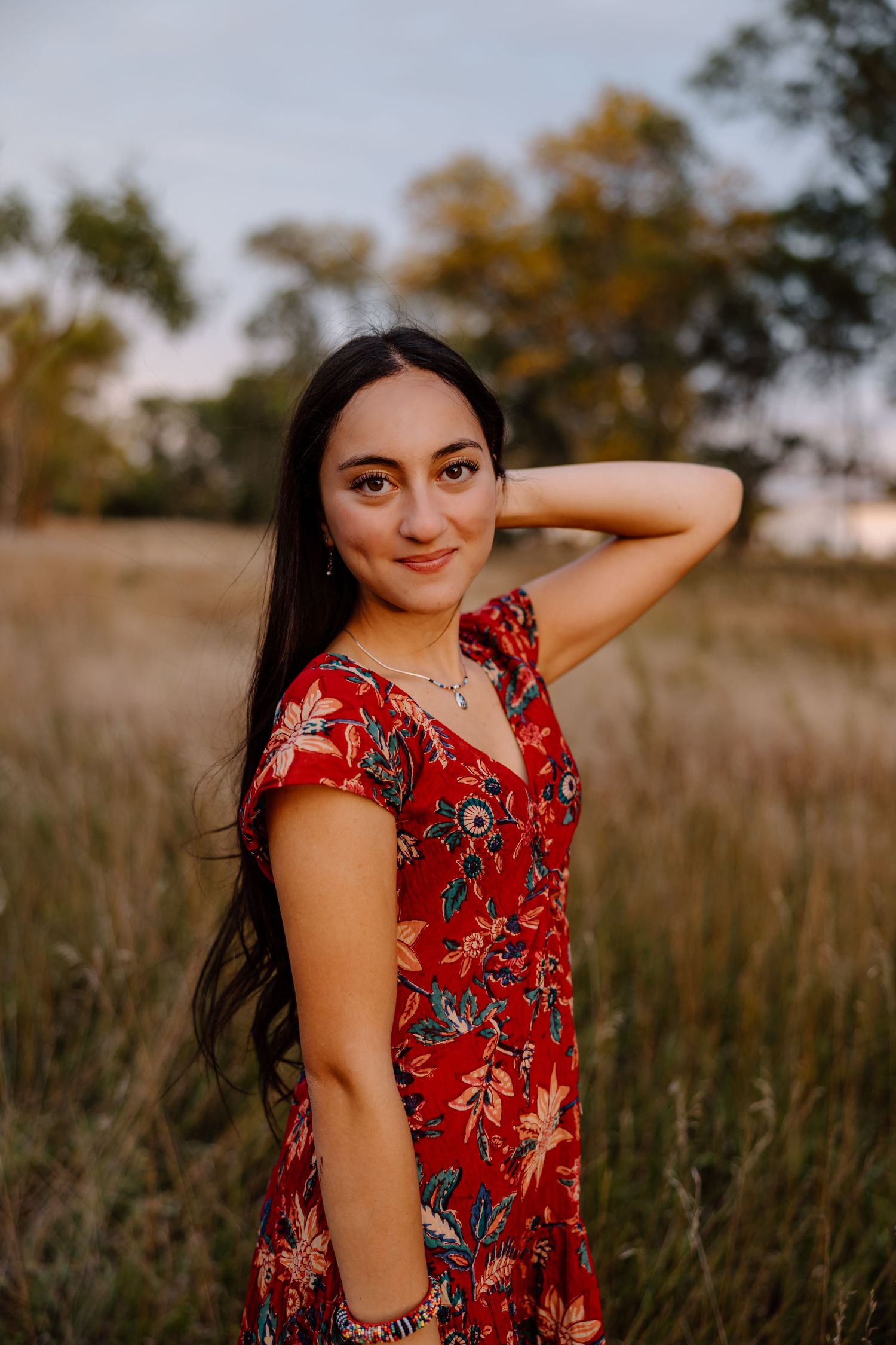 A person in a red floral dress poses in a field at golden hour with tall grass and autumn trees in the background.
