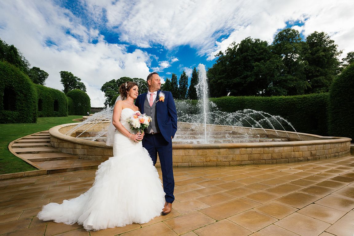 A romantic wedding couple poses in front of elegant garden fountains and stone steps during their outdoor celebration.