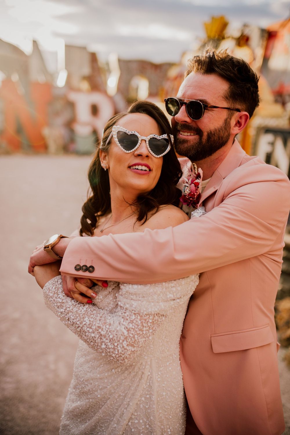 A couple in sunglasses embrace at a wedding, with the bride in white lace and groom wearing a pink suit.