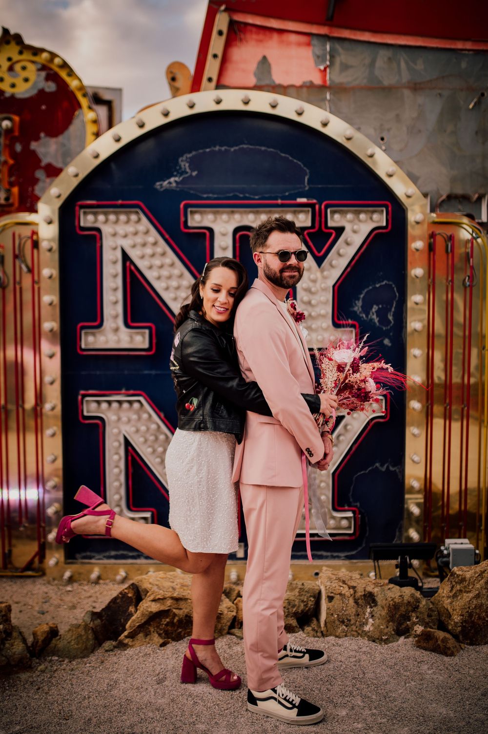 Couple poses against vintage carnival signage backdrop in pink suit and black leather jacket.