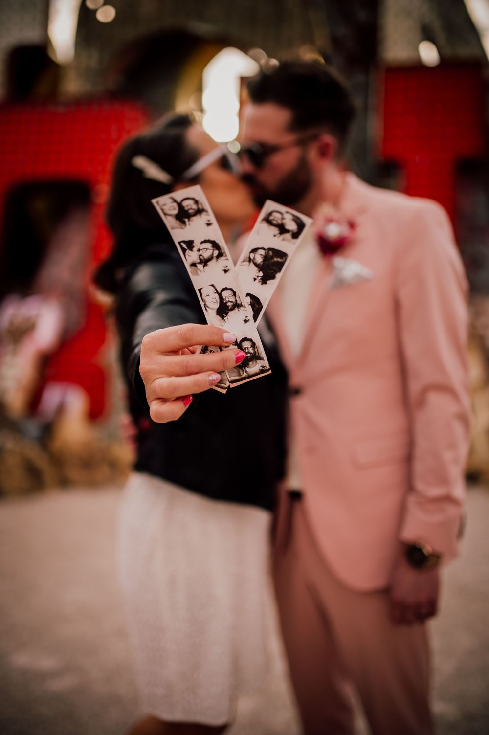 Wedding guests hold vintage photo booth strips while gathered at a reception with pink and red decor.