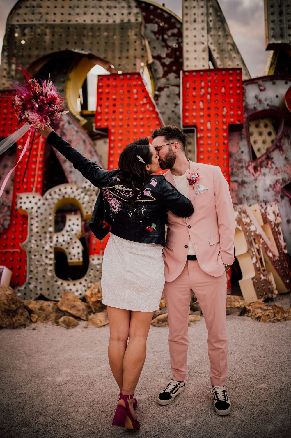 A couple embraces in front of large red letters against an industrial backdrop with decorative lighting.