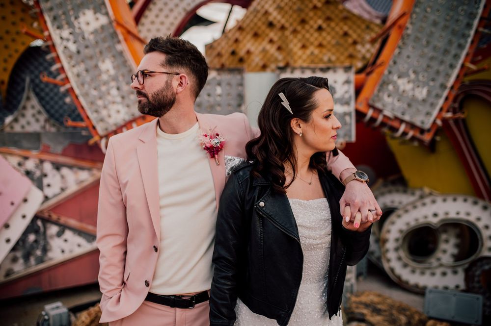 A couple in pink and black outfits poses together at an amusement park with carnival lights in the background.