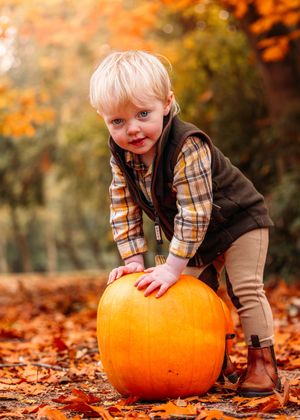 Child playing with large orange pumpkin among fallen autumn leaves.