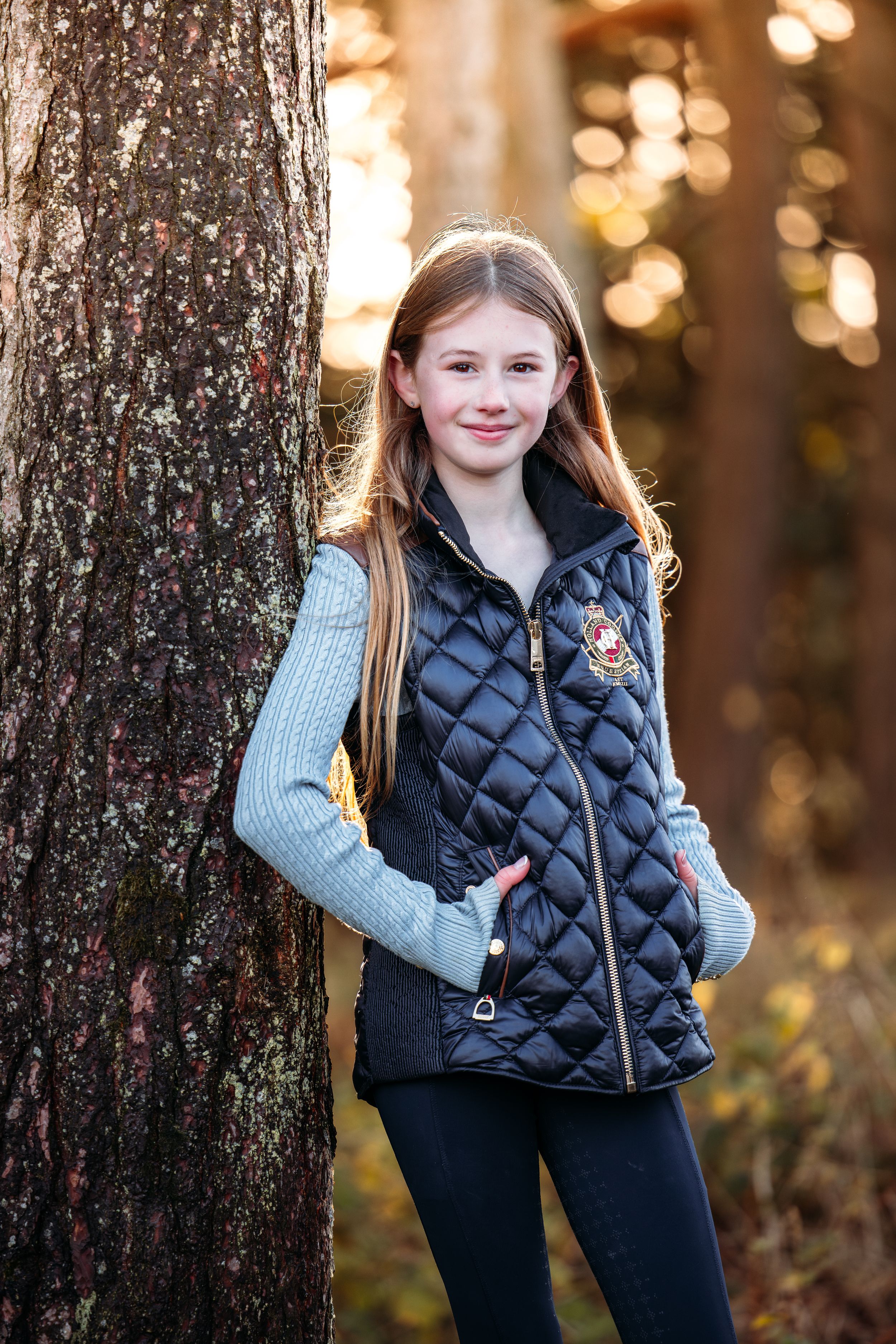 Young person wearing a navy quilted vest and gray sweater poses against tree trunks in autumn.