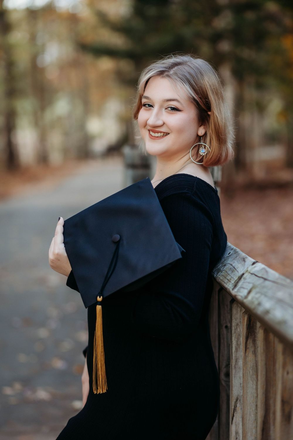 Graduate in black cap and gown with gold tassel poses on wooden railing during autumn.