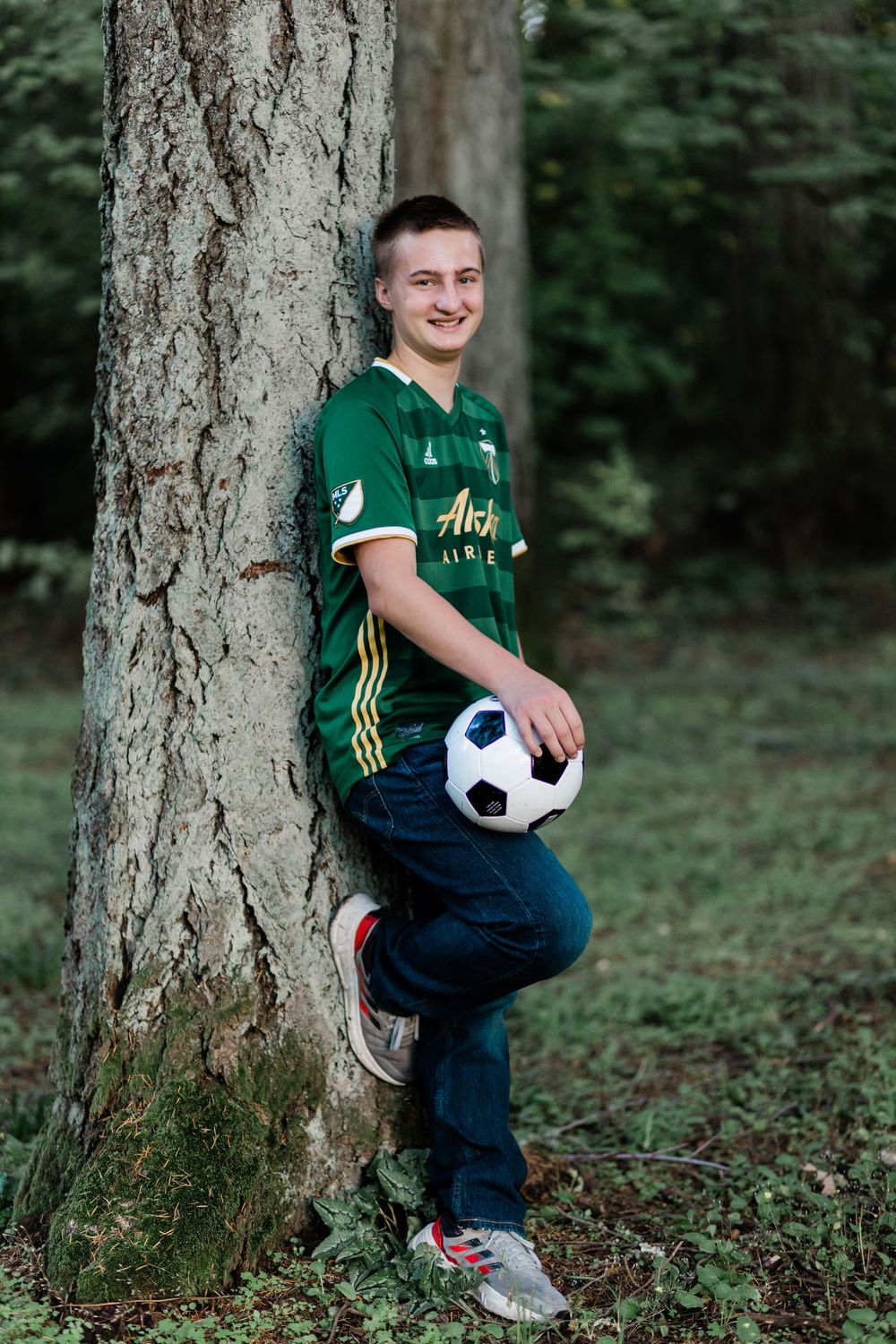 Soccer player in green Portland Timbers jersey poses with ball against large tree trunk.