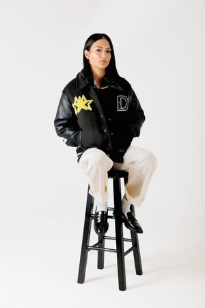 Stylish outfit featuring black varsity jacket and white pants while seated on black stool against white backdrop.