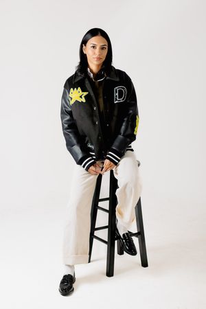 A person wearing a black varsity jacket and white pants poses on a black stool against a white backdrop.