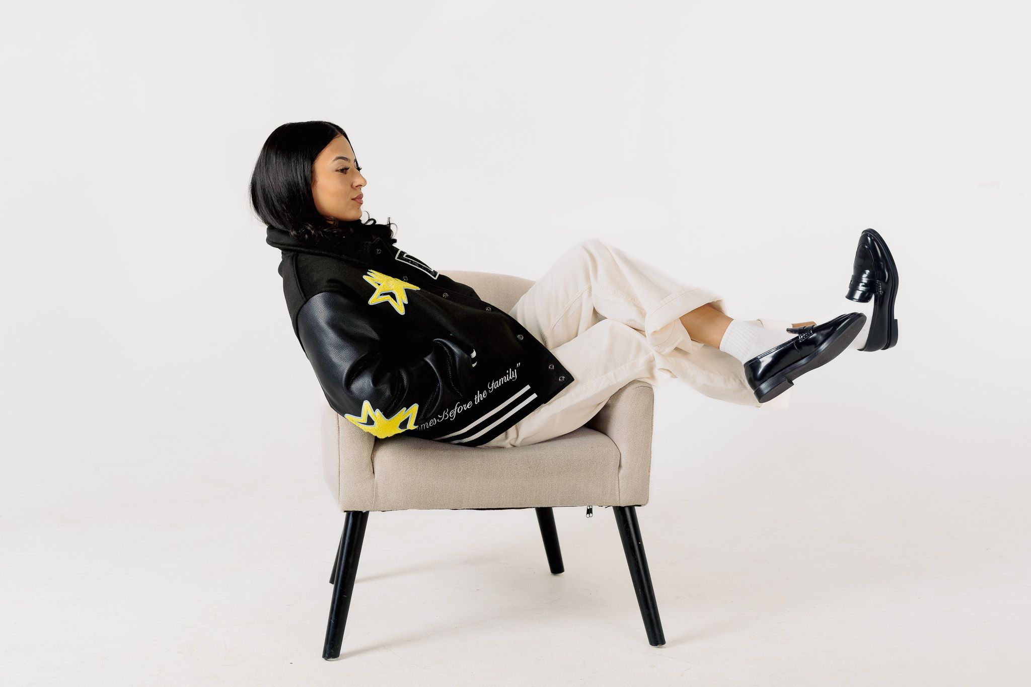 Casual pose in black varsity jacket and white pants while lounging in modern accent chair.