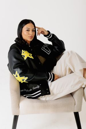 A person in a black varsity jacket with yellow star patches sits casually on a light-colored armchair.