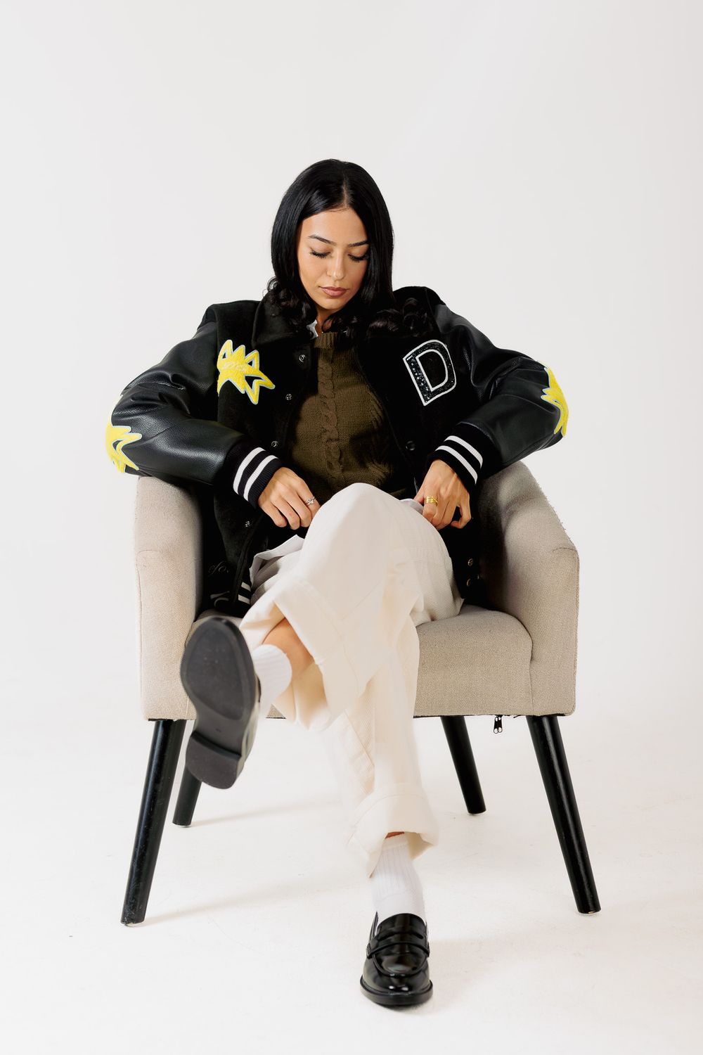 A person in a black jacket and white pants sits casually on a modern gray armchair against a white backdrop.