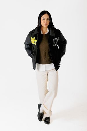 Fashion model wearing black varsity jacket with yellow patches, brown sweater and white pants against white background.