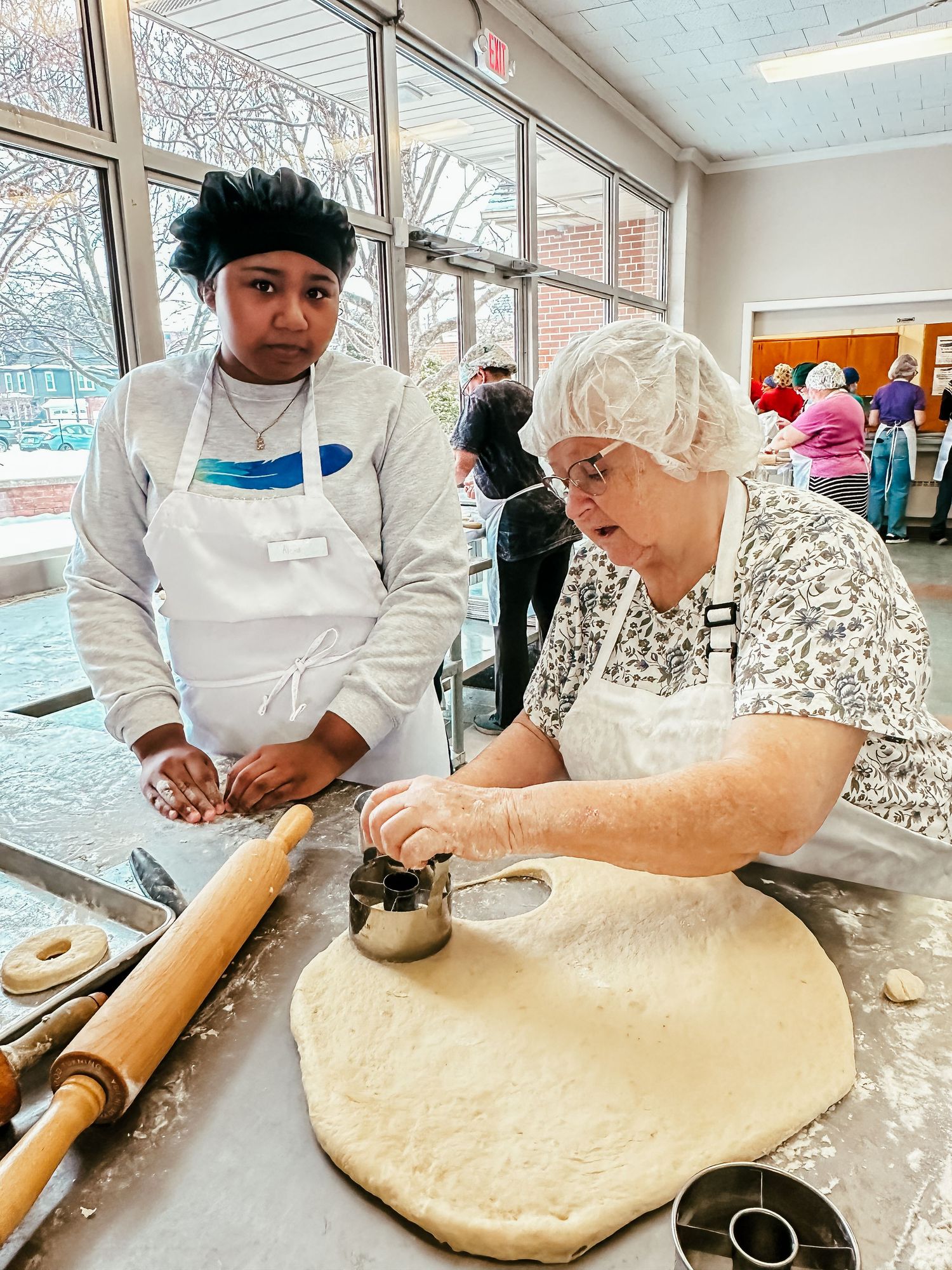 Two people work together on rolling out dough with a rolling pin and flour on a table in a community center.