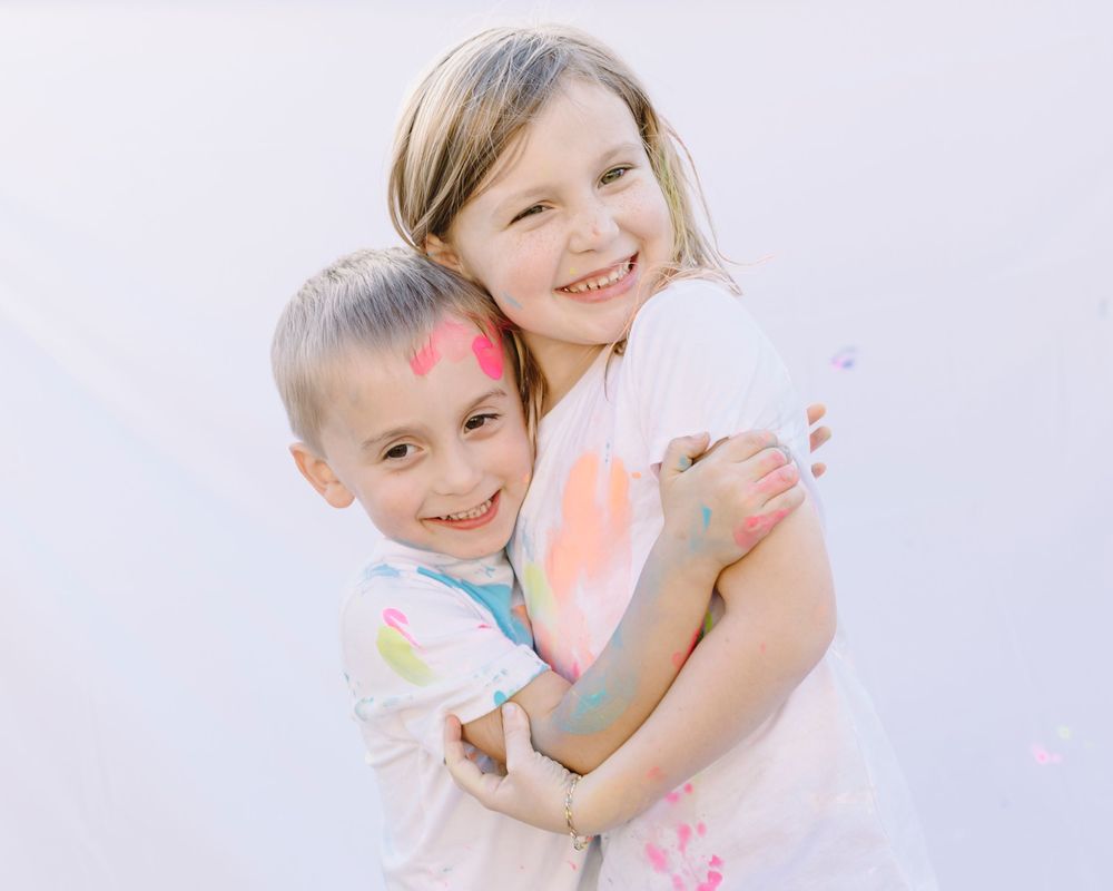 Two children embrace and laugh while covered in colorful powder during a festive celebration.
