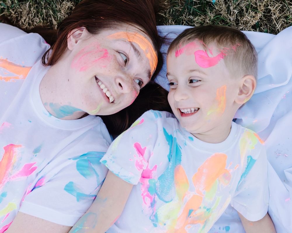 Two people lying down with colorful paint powder on their faces and white shirts smiling at each other.