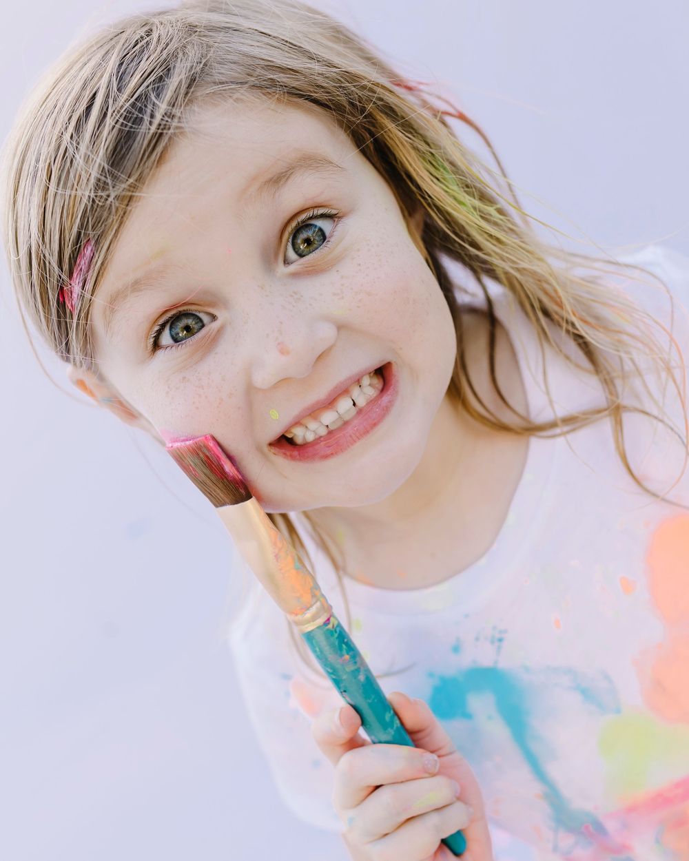 A young child in a colorful setting shows off a bright smile while holding a turquoise marker.