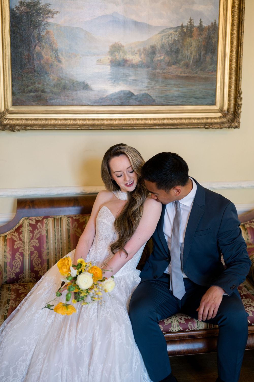 Bride and groom sitting on ornate sofa, bride holding yellow bouquet, groom leaning in close, elegant painting backdrop.