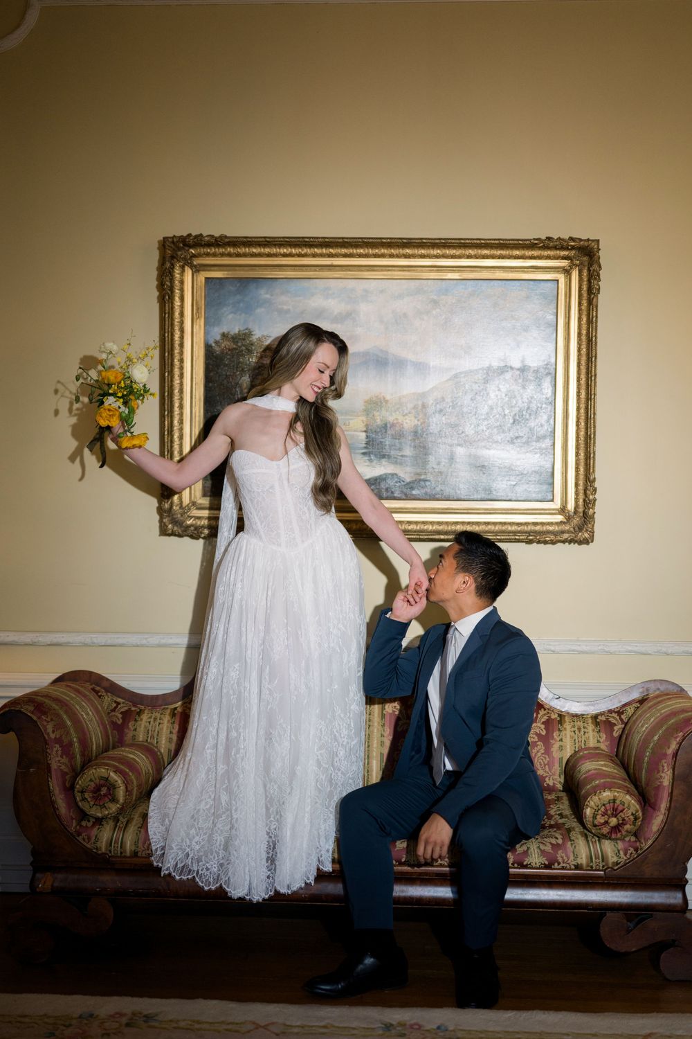 A bride stands on a floral sofa holding yellow flowers while a groom in navy suit kisses her hand before a gold-framed painting.