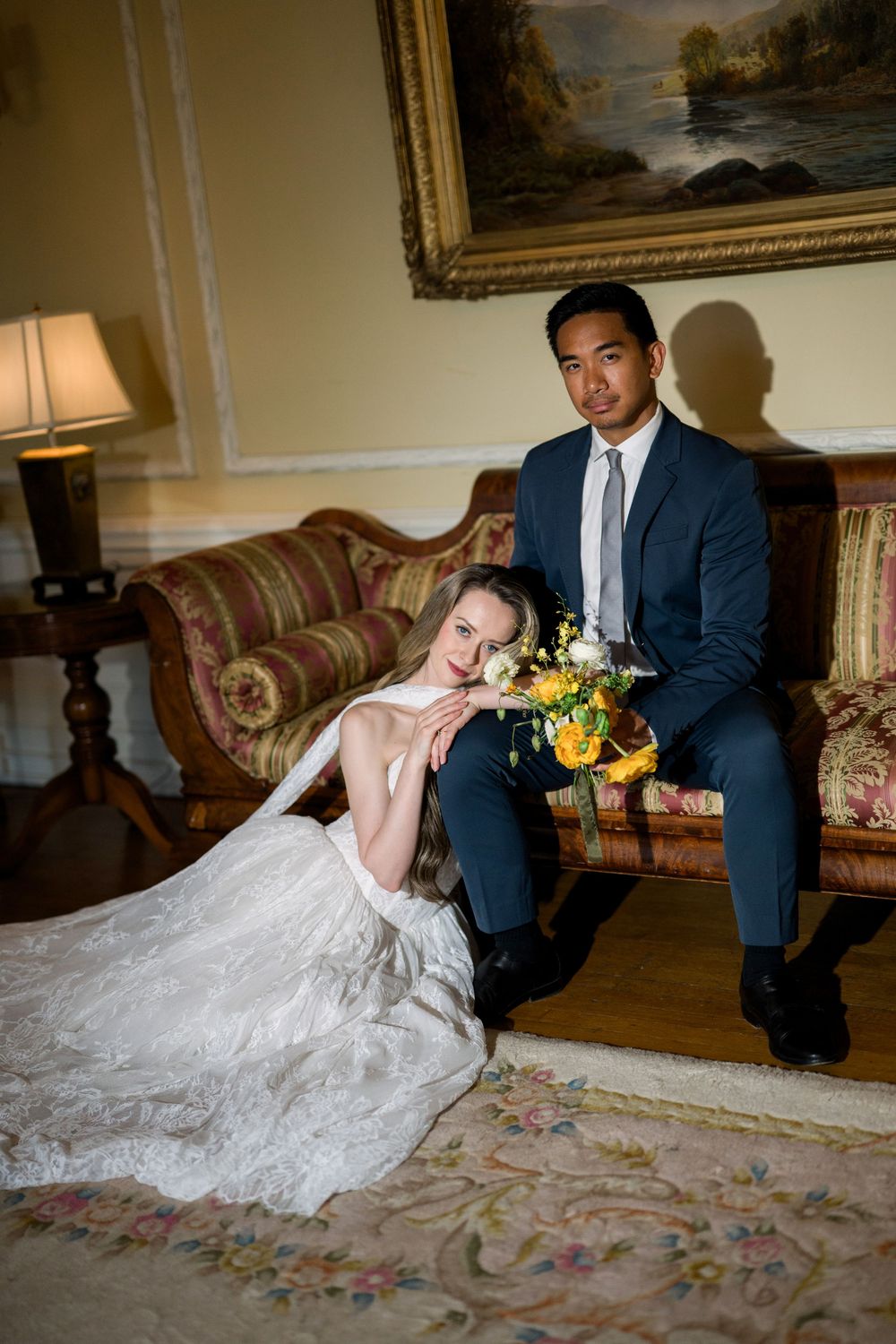 Bride in white gown sits on floor leaning on groom in navy suit on ornate sofa, holding yellow floral bouquet.