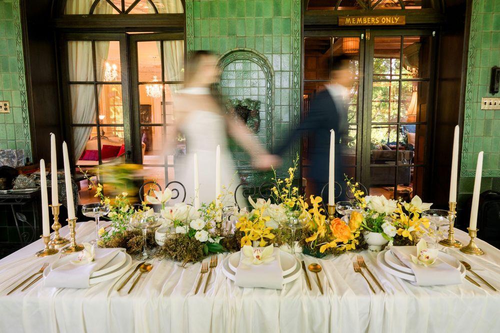 Elegant wedding reception table with gold candles, yellow florals, and a blurred couple holding hands in a green-tiled venue.