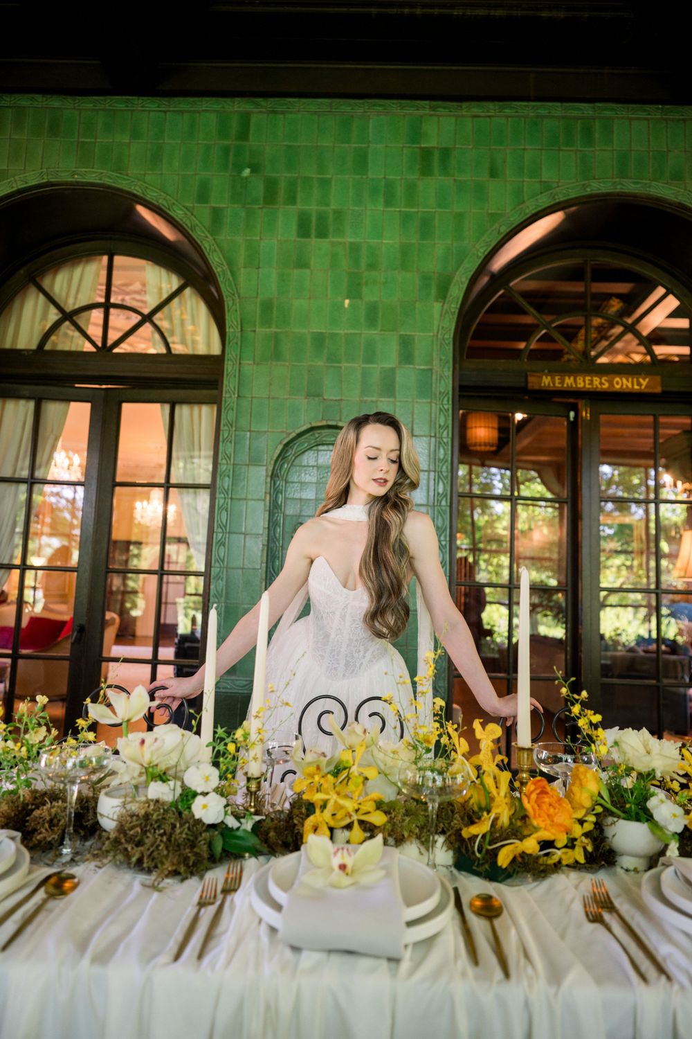 Bride in white gown stands at elegantly decorated wedding table with yellow florals, candles, and gold accents in green-tiled venue.