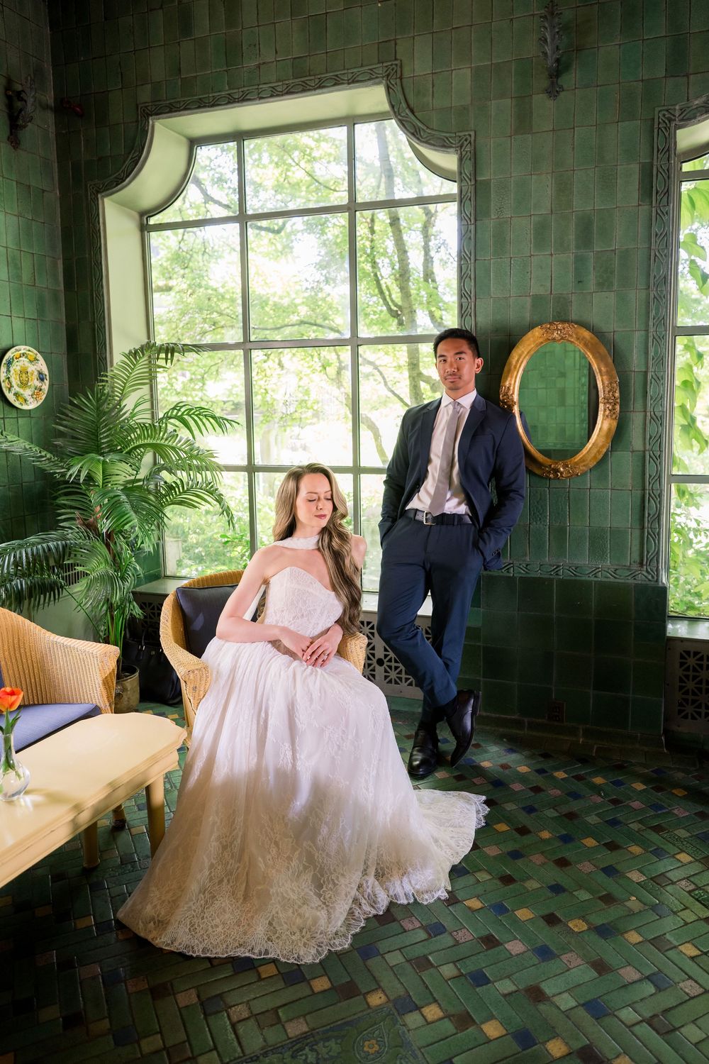 Bride in white lace gown seated beside groom in navy suit in elegant green-tiled room with arched windows.