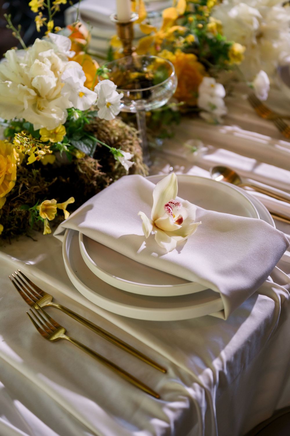 Elegant wedding table setting with gold cutlery, white plates, orchid napkin, and yellow floral centerpiece.